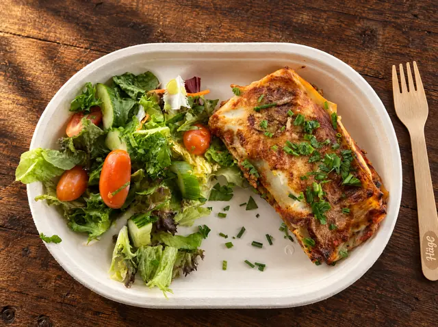 Lasagne mit goldener Käsekruste und Salatmix in der Pappschale auf Holz, daneben eine Holzgabel mit Häge-Logo